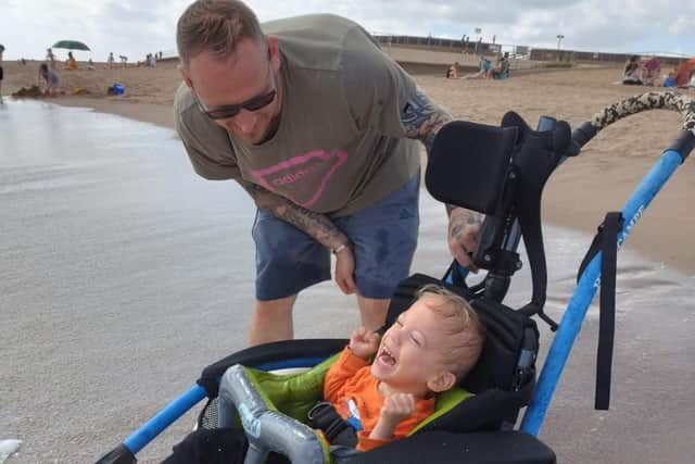 Joey laughing as he is able to go in the sea for the first time, watched by dad Tom.