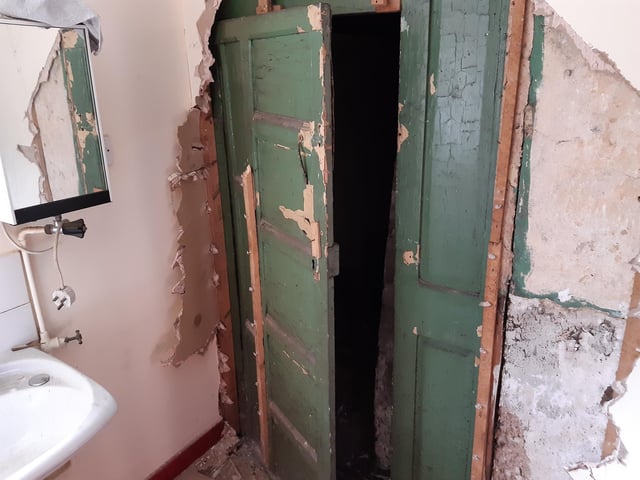 This discovery was made on Halloween and is appropriately spooky. Shane was investigating an unusual ceiling line in the adjacent room. Behind a wall, he found a lockable door. Behind the door (which has wallpaper on the back of it), he found a dark corridor with hooks on the side. At the end of the corridor, he found a small room. That room has one window, high up; however, it is sealed - it also has bars on it. The window appears to line up with one of the staircase landings, but it is covered up from that side.