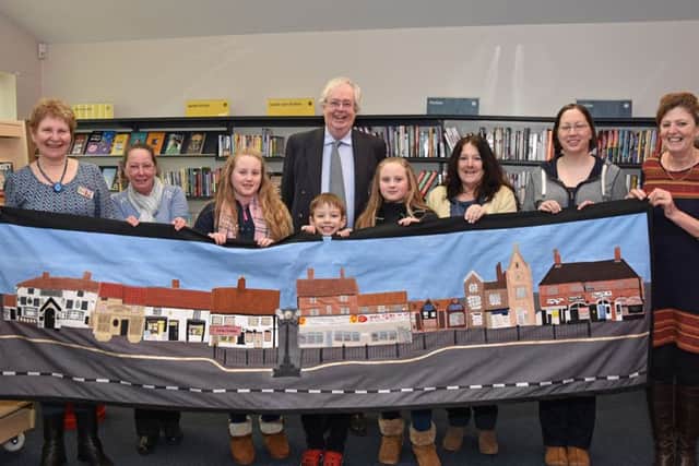 Pictured holding a tapestry of the shops in Wragby Market Place are children, mums and organisers of the recently opened Wragby Community Hub - Barbara Barlett, Maureen Thompson, Amy and Lucy Lowman, Mathew Bacon, Coun Nick Guyatt, Vivien Lewis, Dani Bacon and Clare Lee. Photo by John Edwards EMN-160223-151132001